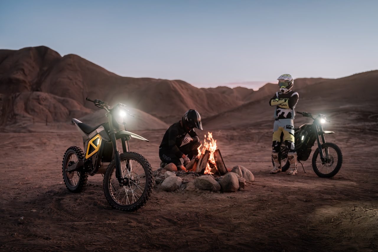 Three riders around a campfire with their bikes at dusk